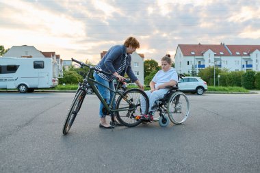 Engelli çocuğun tekerlekli sandalyeden bisiklete geçişi, annenin çocuğa yardım etmesi, aktivite rehabilitasyonu sağlık konsepti.
