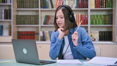 Kulaklıklı kız üniversite öğrencisi bilgisayarla video üzerinden konuşuyor, sınava hazırlanıyor, eğitim binası kütüphanesinde masa başında oturuyor. Video konferansı sohbet çevrimiçi ders webinar