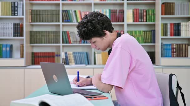 大学図書館にノートパソコンの本を持って座っている若い男子学生 20歳の男性は ヘッドフォンでノートブックを書いています 大学のコンセプト — ストック動画