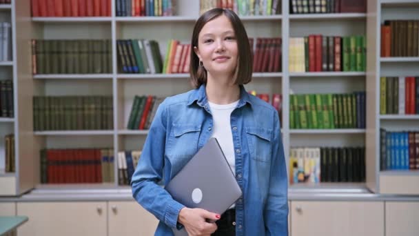 カメラを見ている若い女性大学生の笑顔の肖像画は 教育ビルの図書館教室でラップトップを掲げています 高等教育 若者のコンセプト — ストック動画