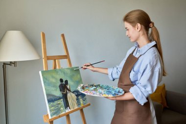 Genç, güzel kadın ressam sehpada akriliklerle resim yapıyor. Genç kız sanat okulu kursları öğrencisi, evde çizim yapıyor. Yaratıcılık hobisi eğitim, sanat terapisi, satılık tablolar, gençlik konsepti