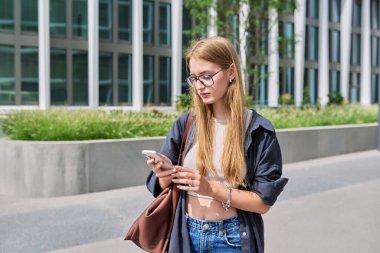 Akıllı telefonu olan genç, ciddi bir kadın, modern şehir, şehir geçmişi. Öğrenci kız cep telefonuna bakıyor, mesajlaşıyor, cep telefonu uygulamalarını iletişim, çalışma ve boş zamanlarında kullanıyor.