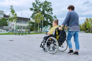 Back view of woman with boy in wheelchair near the school building, copy space. Children, disability, education, activity, health, lifestyle concept clipart