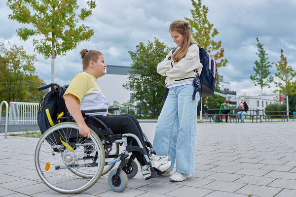 Дети 10, 11 лет друзья одноклассники мальчик в инвалидной коляске и девушка с рюкзаком говорить весело рядом со школьным зданием. Образование, дружба, общение, инвалидность, инклюзивность