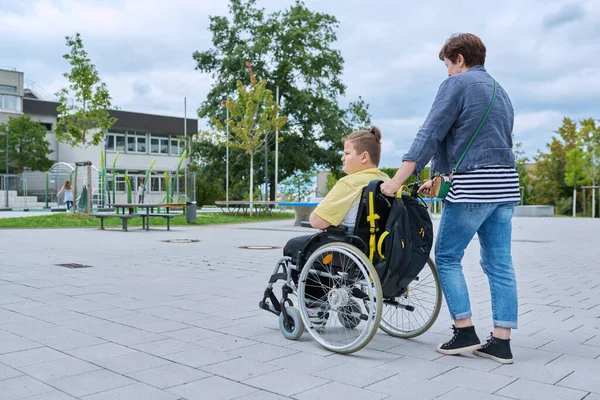 stock image Back view of woman with boy in wheelchair near the school building, copy space. Children, disability, education, activity, health, lifestyle concept