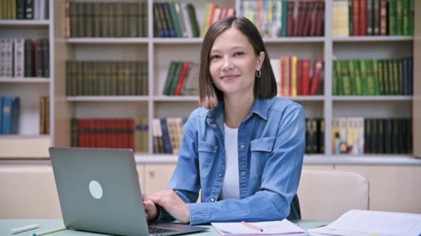 若い女性大学生の笑顔の肖像画は 教育ビルの図書館教室でラップトップを持って机に座っているカメラを見ている 高等教育 若者のコンセプト — ストック動画