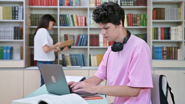 在大学图书馆里 年轻的男生坐在电脑前看书 20岁的家伙 带着耳机在笔记本电脑上打字 大学概念 — 图库视频影像