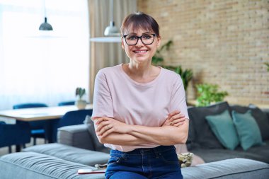 Evde kendinden emin orta yaşlı bir kadının portresi. Kollarını kavuşturup gülümseyen başarılı kadın kameraya bakıyor, oturma odası arka planı.