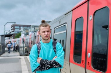 Şehir tren istasyonunda elektrikli tren bekleyen genç bir erkek. 19 yaşında, 20 yaşında, sırt çantalı ciddi bir hippi öğrenci. Şehir ulaşımı, banliyö treni, yolcu taşımacılığı, gençlik