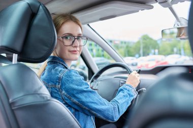 Araba direksiyonunda oturan, kameraya bakıp gülümseyen gözlüklü genç kız sürücü, genç bir kadının araba kullanma ehliyeti var. Gençlik, araba okulu, yaşam tarzı konsepti.