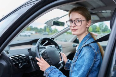 Araba direksiyonunda oturan, kameraya bakıp gülümseyen gözlüklü genç kız sürücü, genç bir kadının araba kullanma ehliyeti var. Gençlik, araba okulu, yaşam tarzı konsepti.