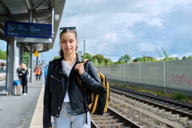 Şehir tren istasyonunda elektrikli tren bekleyen genç bir bayan. Sırt çantalı gülümseyen liseli kız. Şehir ulaşımı, banliyö treni, yolcu taşımacılığı, gençlik