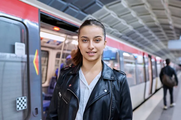 Teenage girl on platform of subway station, against backdrop of subway train, smiling beautiful female looking at camera. Student teenager youth, lifestyle in city, subway passengers, urban transport