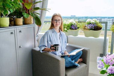 Kitap okuyan, evinde, balkondaki koltukta oturmuş kahve içen genç bir kadın. Rahatlık, yaşam tarzı, rahatlama, okuma, gençlik kavramı