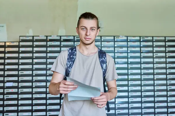 Young guy student reading an important mail letter near mailboxes in student dormitory