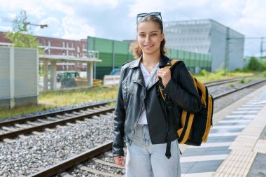 Şehir tren istasyonunda elektrikli tren bekleyen genç bir bayan. Sırt çantalı gülümseyen liseli kız. Şehir ulaşımı, banliyö treni, yolcu taşımacılığı, gençlik