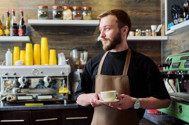Önlüklü genç barista erkek portresi. Yeni hazırlanmış cappuccino kahvesi. Kahve dükkanında tezgahın arkasında duruyor. Küçük işletme hizmeti, kafe, personel iş konsepti