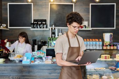 Kafeteryada dizüstü bilgisayarı olan önlüklü genç bir adam. Gençlik, iş, çevrimiçi teknoloji sipariş alıyor
