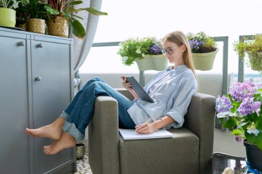 Liseli kız evde ders çalışıyor açık terasta koltukta rahatça oturuyor ev bitkileriyle birlikte dijital tablet kullanarak not defterine not alıyor. Eğitim teknolojisi ergenlik çağı