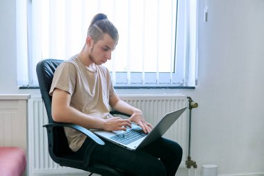 19 yaşında, 20 yaşında, genç bir öğrenci evinde oturmuş bilgisayar laptopunu kullanıyor, okuyor ve daktilo ediyor. İletişim eğlence işleri, eğitim ve gençlik kavramlarını öğrenmek için dijital teknolojiler