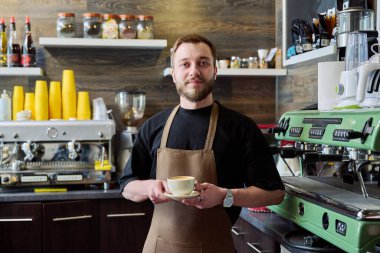 Önlüklü genç barista erkek portresi. Yeni hazırlanmış cappuccino kahvesi. Kahve dükkanında tezgahın arkasında duruyor. Küçük işletme hizmeti, kafe, personel iş konsepti