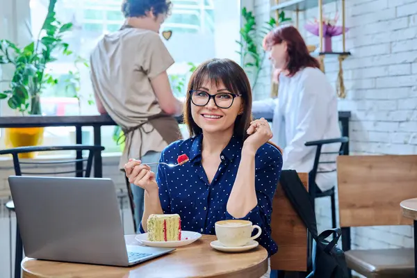 Middle aged woman in bakery cafeteria with cup of coffee and dessert cake, sitting at table, with laptop, happy smiling female looking at camera. Lunch break, food, lifestyle, mature 40s people