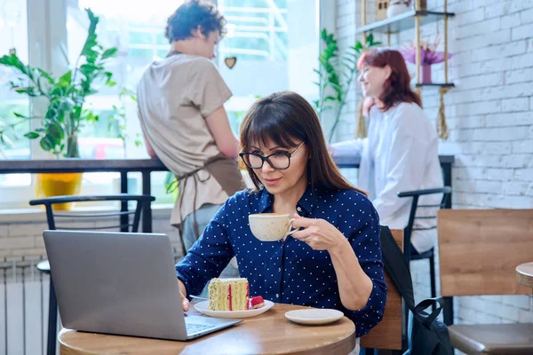 Middle aged woman in bakery cafeteria with cup of coffee and dessert cake, sitting at table, with laptop. Lunch break, food, lifestyle, mature 40s people concept
