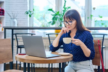 Orta yaşlı bir kadın, kahve içiyor, laptop ekranına bakıyor, kafede masada oturuyor, kahve dükkanında. Uzaktan çalışma, serbest çalışma, blog yazma, sohbet, online internet teknolojisi, olgun insanlar konsepti