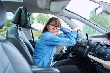Orta yaşlı, gözlüklü, direksiyonda oturan, kameraya gülümseyen bir kadın sürücü. Ulaşım, yaşam tarzı, sürüş, sigorta onarım araba bakımı, olgun insanlar konsepti
