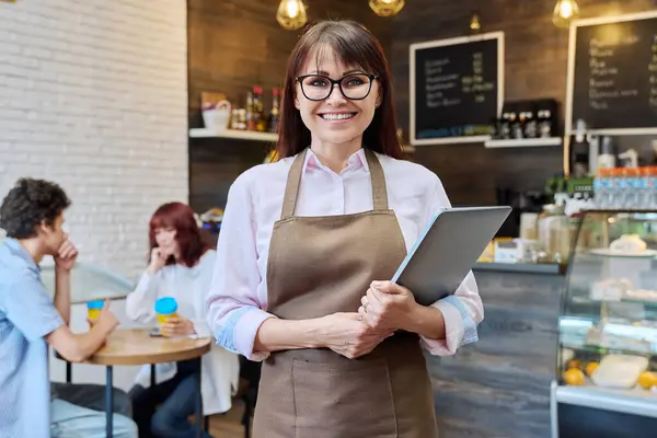 Portrait of middle aged woman owner of bakery coffee shop worker, smiling looking at camera female with laptop in her hands, inside cafeteria. Small business, entrepreneur, work, people concept