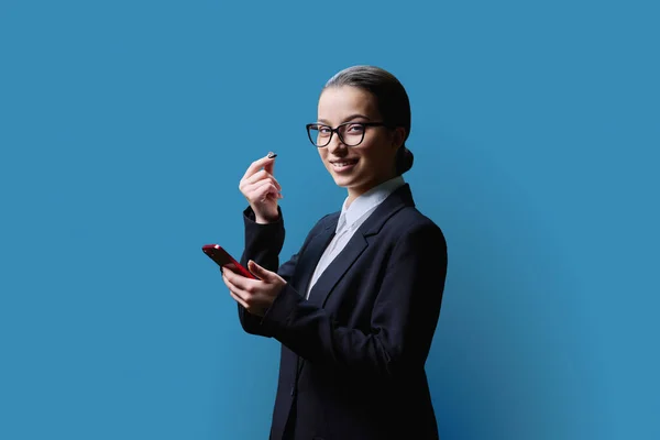 Young teenage girl making money gesture, rubbing her fingers, looking at camera with smartphone in her hands, on blue color studio background. Body language, finance, money for teenage students