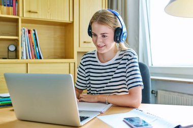 Kulaklıklı kız lise öğrencisi bilgisayarda dizüstü bilgisayar kullanıyor. Evde uzaktan eğitim gören bir genç test ve teknolojik ergenlik için hazırlanan eğitim web sitelerini dinliyor.