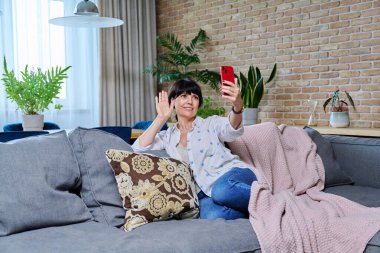 Oturma odasındaki koltukta oturan olgun, rahat bir kadın telefon konferansı için akıllı telefonu kullanarak telefon ekranına el sallıyor. Teknoloji yaşam tarzı boş zaman iletişimi 50 kişi