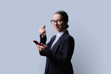 Genç kız para jesti yapıyor, parmaklarını ovuşturuyor, elinde akıllı telefonuyla kameraya bakıyor, gri renkli stüdyo arka planında. Vücut dili, finans, genç öğrenciler için para