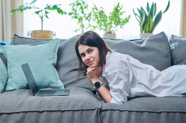 Genç bir kadın dizüstü bilgisayarla kanepede uzanıyor, ev içi oturma odası. Yaşam tarzı, boş zaman, insanların konsepti