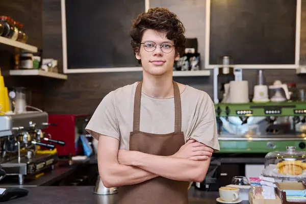 Portrait of young confident male coffee shop worker, with crossed arms, in apron, looking at camera, coffee machine background. Youth, work, employee, staff concept
