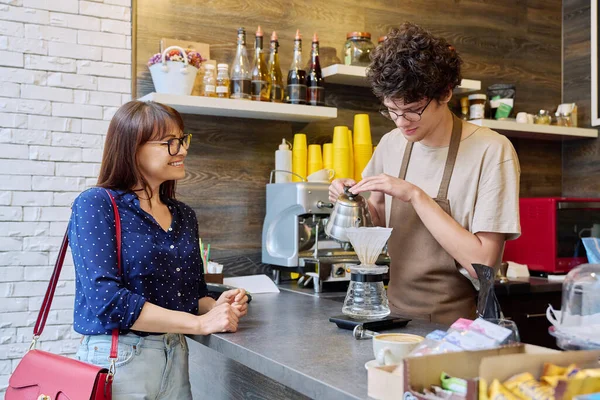 Young male barista talking to woman customer in coffee shop, near counter with cup of freshly prepared cappuccino coffee. Small business, food service occupation, staff, work concept