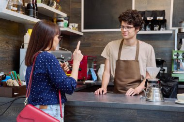 Genç erkek barista kahve dükkanında kadın müşteriyle konuşuyor, tezgahın yanında taze hazırlanmış cappuccino kahvesiyle. Küçük işletme, yemek servisi işi, personel, iş konsepti