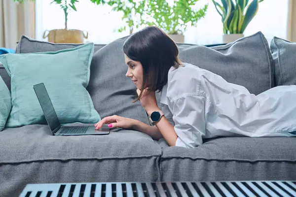 stock image Young woman typing on laptop while lying on sofa at home. Serious female using computer for leisure work study communication, social media, blog, freelancing, internet technology, people concept