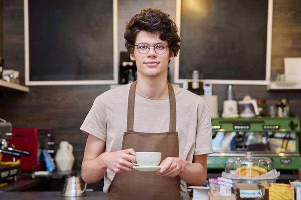 Young guy coffee shop worker, waiter in an apron with cup of coffee, smiling looking at camera inside cafeteria. Youth, work, employee, staff concept
