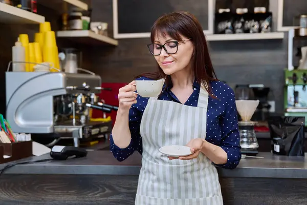 Portrait of successful female small business owner holding fresh cup of coffee, near coffee shop counter. Smiling middle aged woman waiter, worker in apron. Food service occupation, staff, job, work