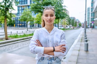 Modern şehrin caddesinde çapraz kollarıyla kameraya bakan kendine güvenen genç kızın portresi. Ergenlik, kentsel yaşam tarzı, gençlik kavramı