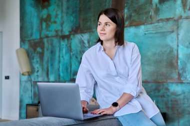 Kanepede dizüstü bilgisayarıyla oturan genç bir kadın, ev içi oturma odası. Yaşam tarzı, boş zaman, insanların konsepti