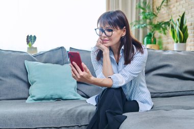 Oturma odasındaki koltukta oturan akıllı telefonlu orta yaşlı bir kadın. Zarif, olgun bir kadın cep telefonu, mobil uygulamalar, online internet teknolojisi, iş olanakları iletişimi kullanıyor.