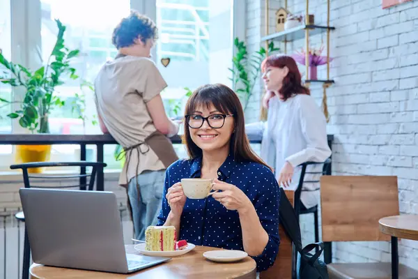 Middle aged woman in bakery cafeteria with cup of coffee and dessert cake, sitting at table, with laptop, happy smiling female looking at camera. Lunch break, food, lifestyle, mature 40s people