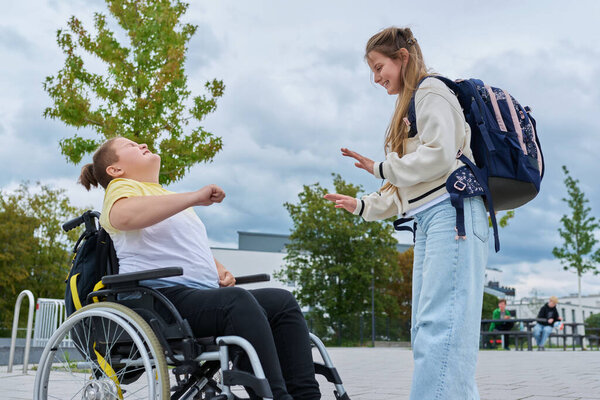 Дети 10, 11 лет друзья одноклассники мальчик в инвалидной коляске и девушка с рюкзаком говорить весело рядом со школьным зданием. Образование, дружба, общение, инвалидность, инклюзивность