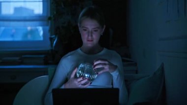 Genç, heyecanlı bir bayan dizüstü bilgisayarda film seyrediyor, evdeki kanepede oturuyor, akşam ışığında. Rahatlama, rahatlama, teknoloji, gençlik kavramı