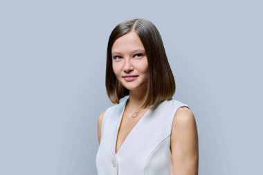 Gri stüdyo arka planında zarif bir iş kadınının portresi. Kameraya beyaz klasik yelekli ve doğal makyajlı başarılı bir kadın bakıyor. Gençlik güzellik sektörü eğitimcileri