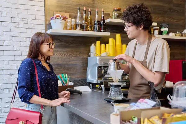 Young male barista talking to woman customer in coffee shop, near counter with cup of freshly prepared cappuccino coffee. Small business, food service occupation, staff, work concept