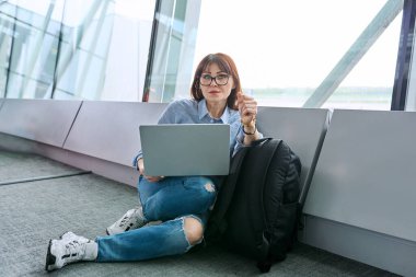 Sırt çantalı orta yaşlı bir kadın havaalanı terminalinde uçak bekliyor, dizüstü bilgisayar kullanıyor, kameraya bakıyor. Seyahat, iş gezisi, yolcu taşımacılığı konsepti